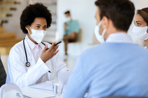 Black female doctor wearing face mask while talking to a couple at clinic.