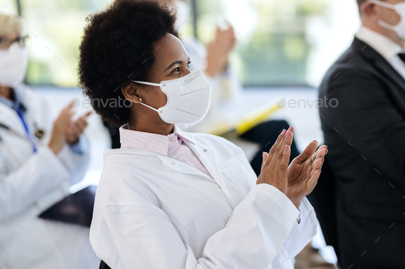 Happy Black doctor wearing face mask and applauding on a seminar in board room.