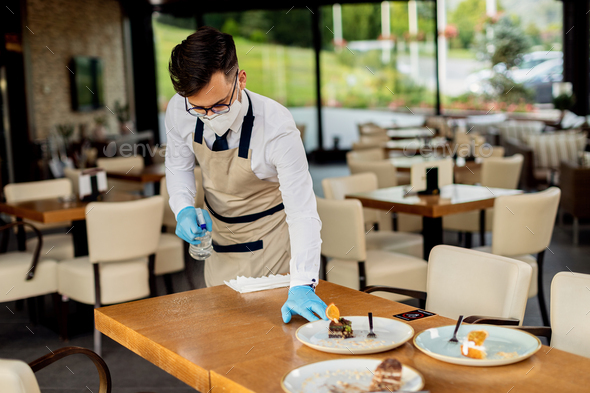 Waiter with protective face mask cleaning table in a cafe.