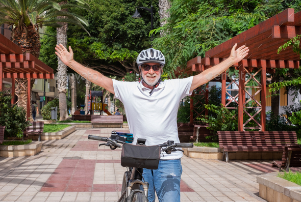 Senior cyclist man wearing helmet standing with open arms in public park looking at camera smiling