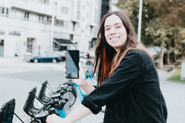 Super happy woman showing phone to camera while renting an electric bike in the middle of the city.