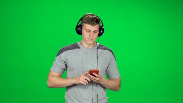 Man in Big Headphones Goes and Texting with Smartphone on Green Screen at Studio alt