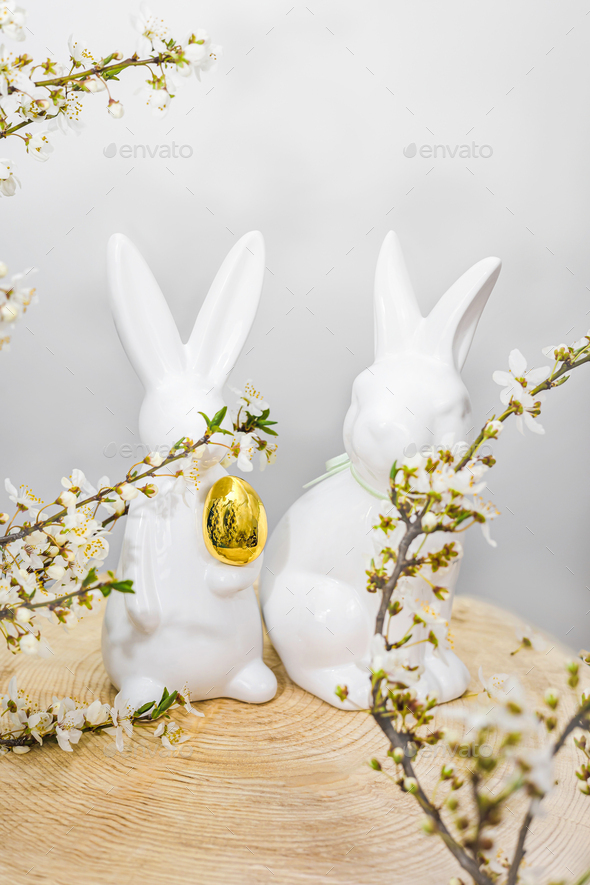 Two white ceramic rabbits on a wooden pallet in blossoming cherry branches with a golden egg.