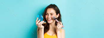 Beauty. Modern girl with candid smile applying make up with brush, looking in pocket mirror