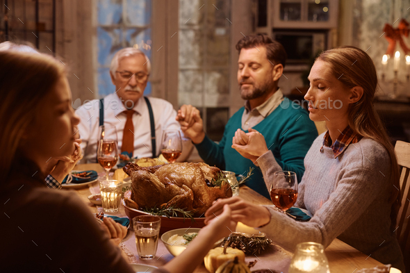 Multigeneration family holding hands while praying during Thanksgiving dinner at dining table.