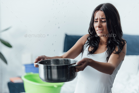 Sad woman holding pot under leaking ceiling in bedroom