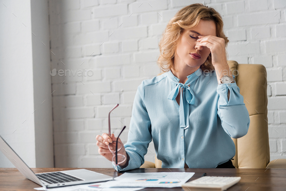 tired businesswoman holding eyeglasses and rubbing nose bridge at workplace