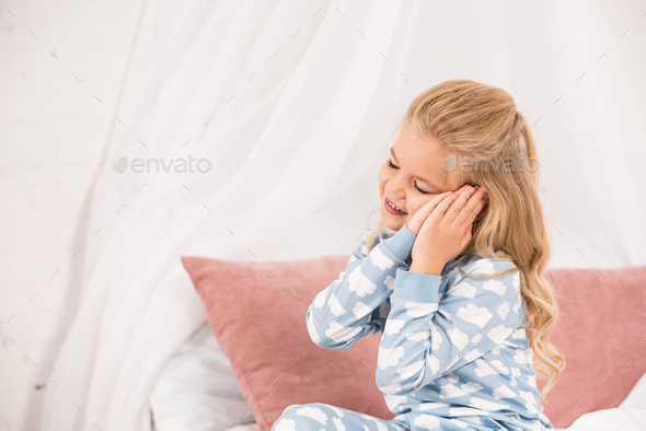 cute sleepy child sitting on bed with closed eyes and hands near ears
