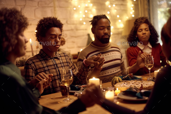 Black boy and his family holding hands while praying during Thanksgiving meal at dining table.