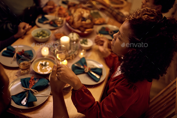 Black woman and her family saying grace while having dinner at dining table on Thanksgiving.