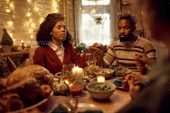 Black couple holding hands and praying while celebrating Thanksgiving with family at dining table.