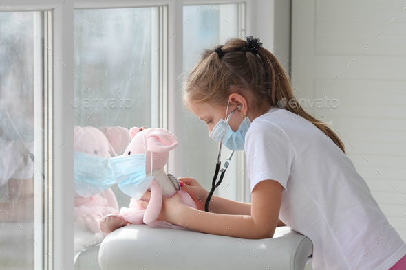 Little girl in medical mask holding stethoscope listening to sick toy, plush rat, patient