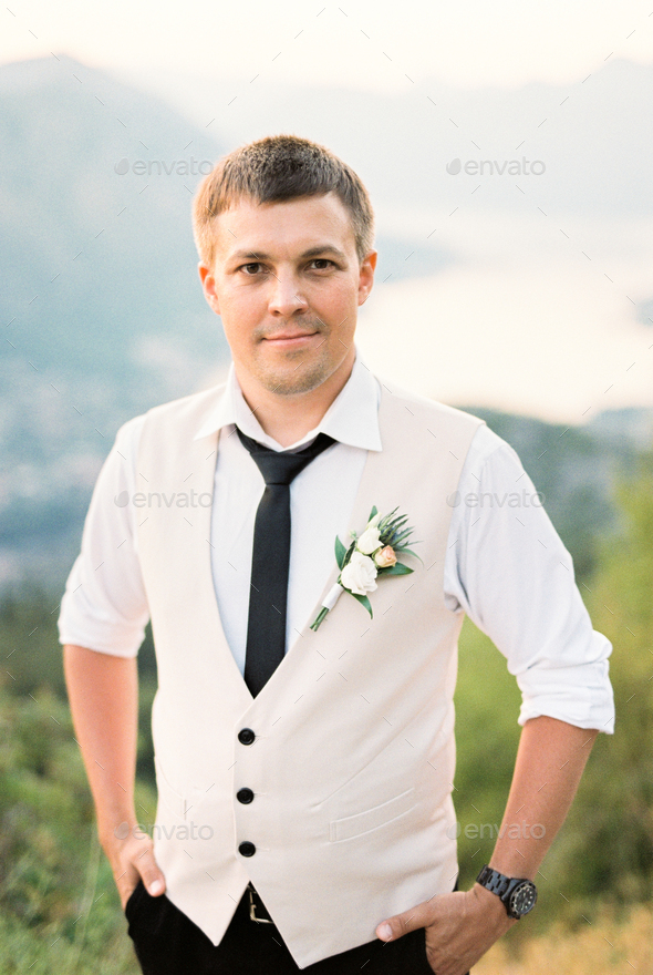 Groom in a vest with a boutonniere stands on a mountain with his hands in his pockets