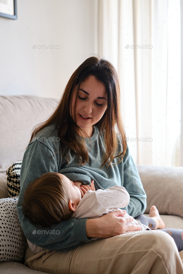 Mother breastfeeding her daughter while sitting on the sofa at home ...