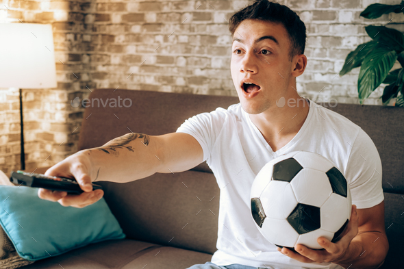 Fan boy watching football on TV holding a soccer ball disappointed for his team\'s missed goal