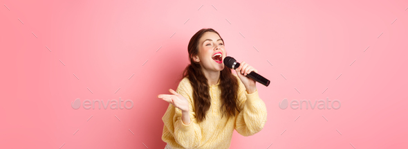 Happy young woman enjoying playing karaoke, singing in mic, looking aside at screen with lyrics