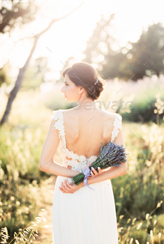 Bride with a bouquet of lavender behind her back stands in a field with her head turned. Back view
