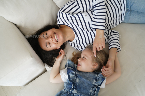 Happy adopted little kid girl tickling laughing mother, lying, having fun together on sofa, top view