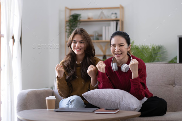 Two Young womanWatching TV Shaking Fists In Joy Celebrating Victory Of Favorite Sport Team Sitting