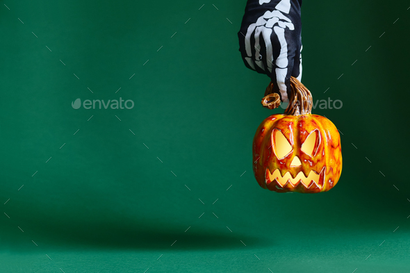 Skeleton gloved hand holds ceramic pumpkin jack-o\'-lantern on green background