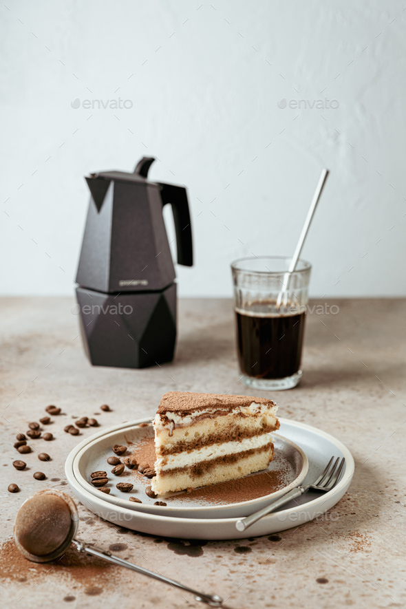 A piece of tiramisu cake served on a plate, a moka pot coffee maker, a glass of black coffee, beans