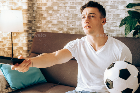 Fan boy watching football on TV holding a soccer ball disappointed for his team\'s missed goal