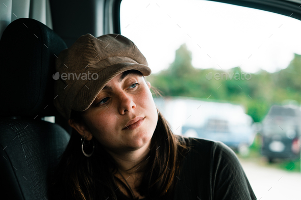 blue-eyed caucasian girl with a hat on her head and big round earrings sitting sideways on a car