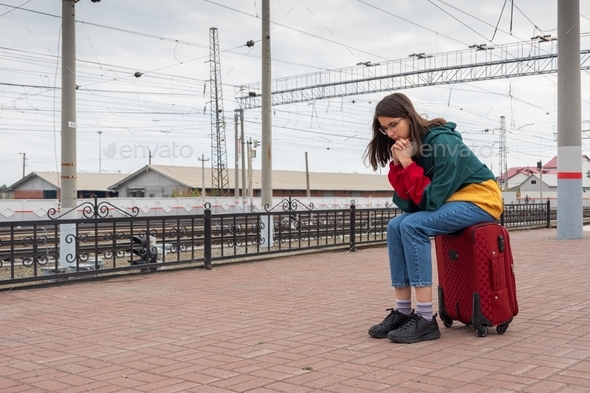 Upset tired girl student in bright clothes sitting on a suitcase at the train station.