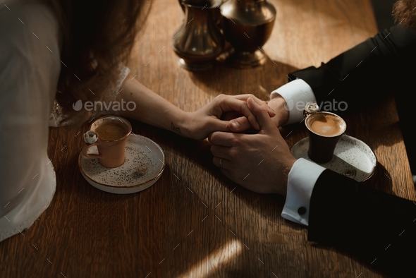 two lovers holding hands while two coffee cups are getting cold on the table