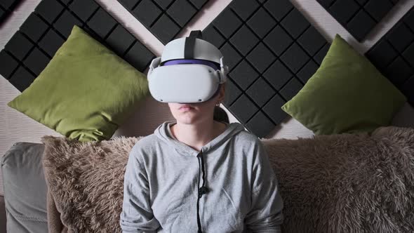 Young Woman in Virtual Reality Helmet Sitting on Sofa and Move Hands at Home alt