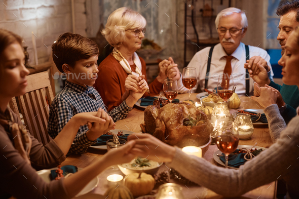 Boy and his extended family holding hands while praying before Thanksgiving dinner at dining table.