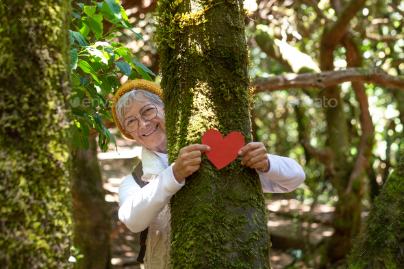 Smiling senior woman partially hidden by a moss covered tree trunk in the woods holding a paper hear