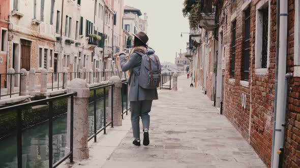 Happy Young Stylish European Woman Tourist with Camera Enjoying Walking Along Famous Old Canal alt