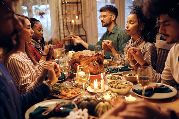 Multiracial group of friends saying grace during Thanksgiving meal at dining table.