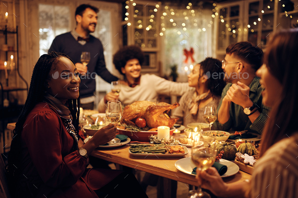 Happy black woman drinks wine and enjoys in conversation with friends during Thanksgiving dinner.