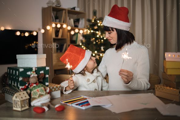 Cheerful cute girl and her mother with charming smile in Santa hats sitting on table with sparkles