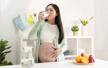 Young Asian pregnant woman drinking water in kitchen room.