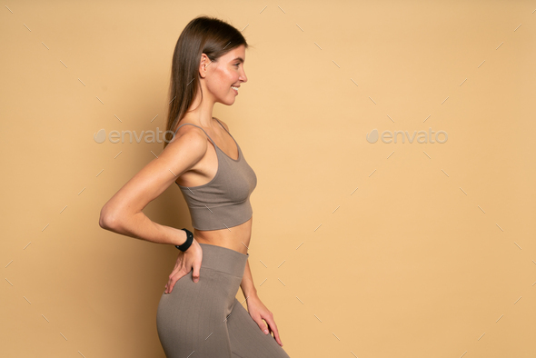 Profile view of athletic woman on brown background with hand on hip showing her flat stomach