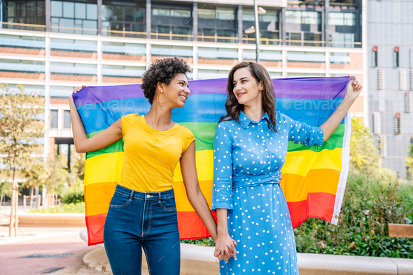 Smiling Multiethnic Lesbian Couple Holding Hands And Raising Rainbow