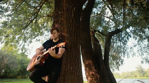 A young guy in a black T-shirt plays an acoustic guitar leaning against a tree.
