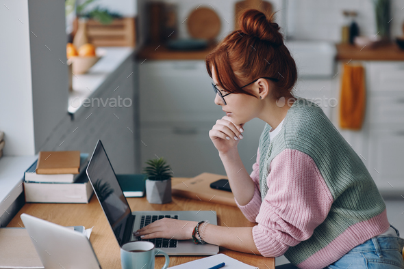 Confident young woman using laptop while sitting at the kitchen counter at home