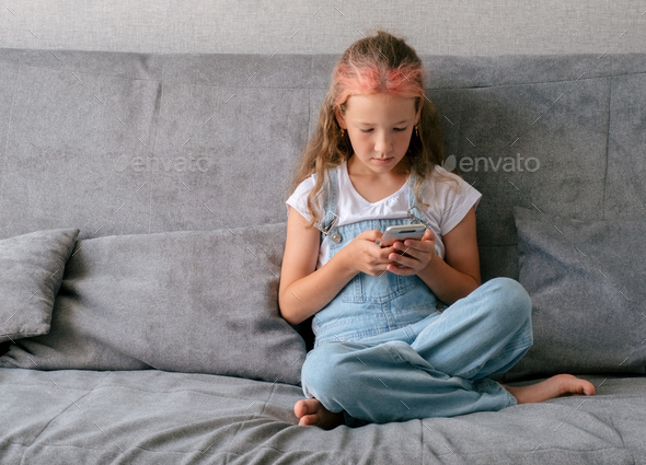 Little girl sitting on couch and using smartphone. Concept of children\'s gadgets addiction.