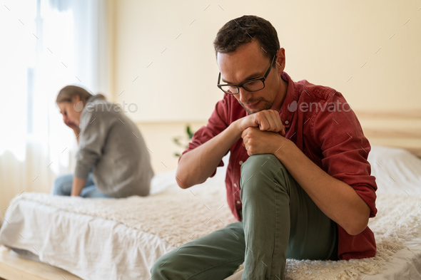 Unhappy Caucasian spouses man and woman sit on different sides of bed in apartment bedroom