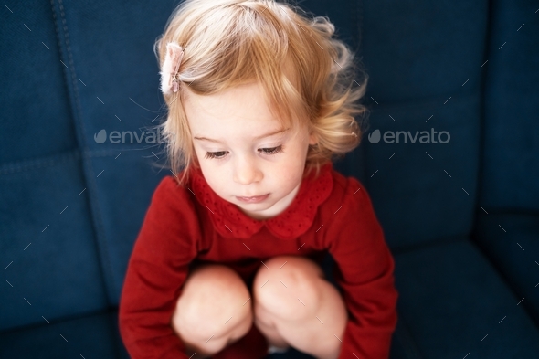 Sad cute caucasian baby girl, toddler with blond curly hair sitting on sofa hugging knees to chest