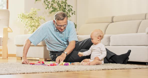 An Elderly Man Dressed in a Tshirt and Smart Pants is Sitting on the Carpet alt