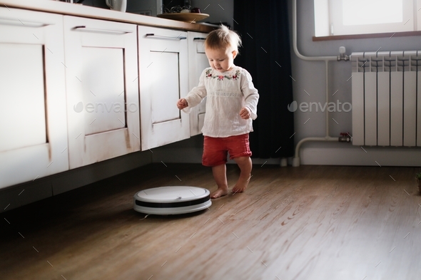 Caucasian baby toddler playing walks behind a robot vacuum cleaner in a stylish apartment