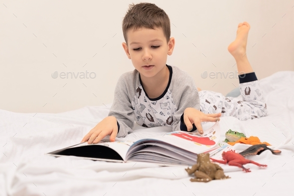 Child boy lies on the bed and reads a book to his toys