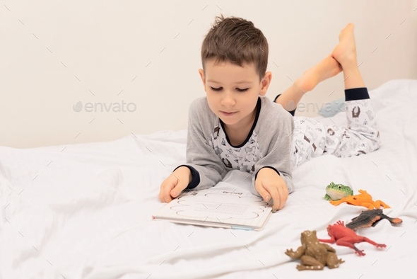 Child boy lies on the bed and reads a book to his toys. Relaxing at home concept.
