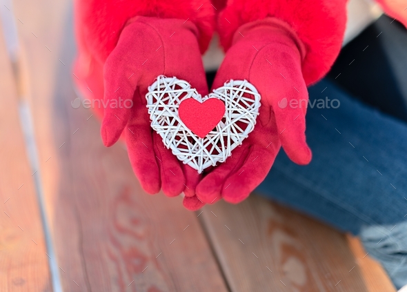 Small red heart on white rattan heart in woman hand in red gloves on wooden background.