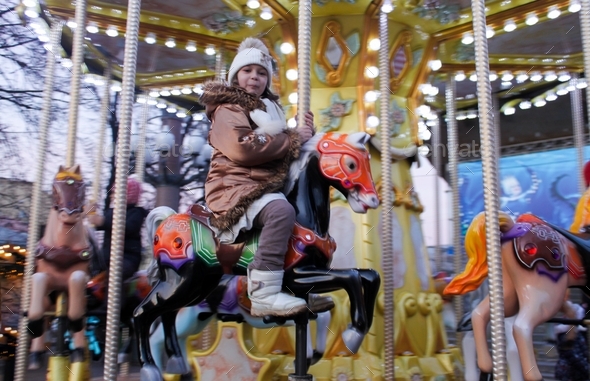 Funny toddler girl in warm clothes rides a merry-go-round horse during the winter Christmas fair